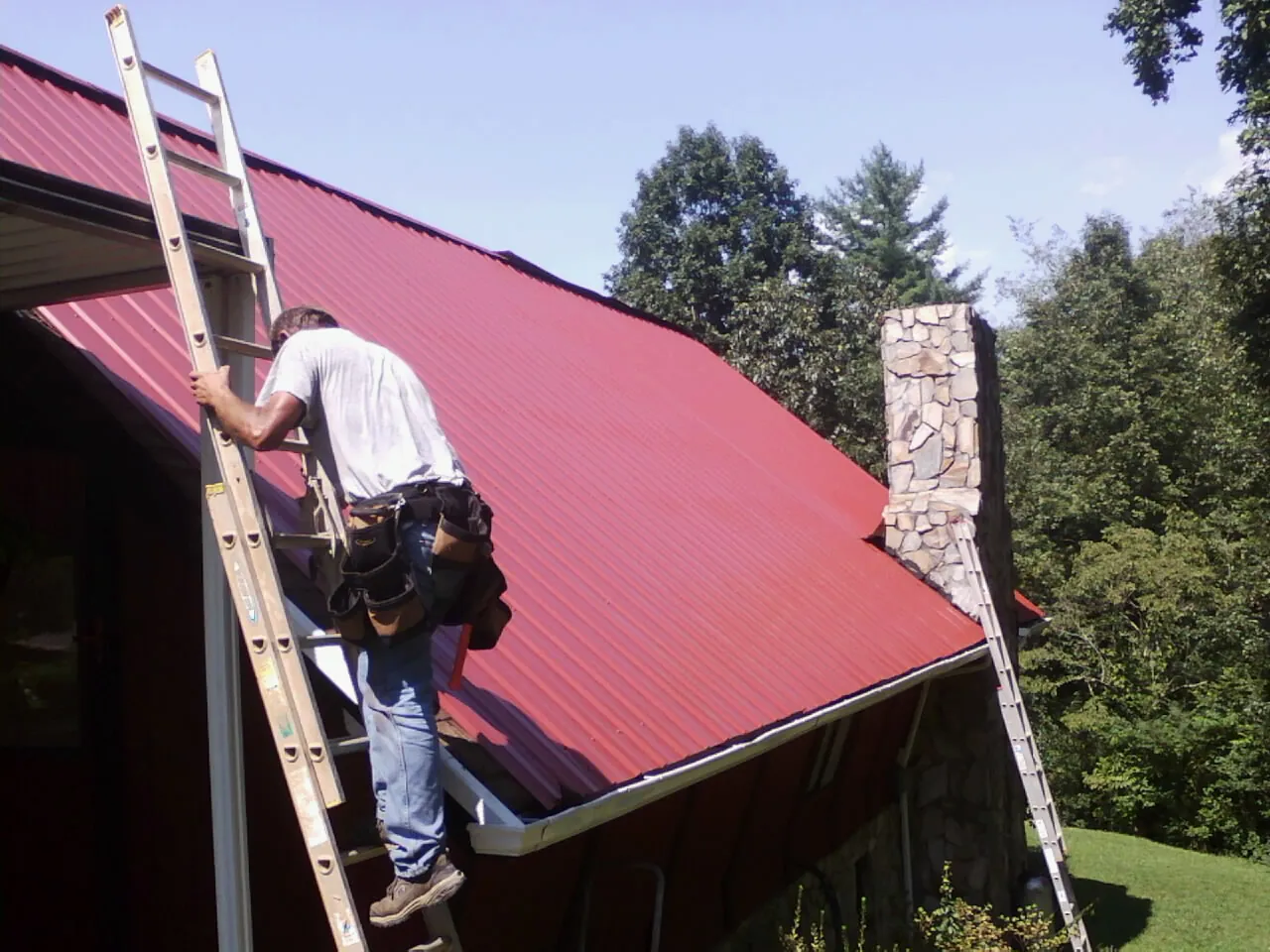 Roofer on a ladder beside a red metal roof and stone chimney — Soffit & Fascia Repair by Roofing Brothers in Jonesburg